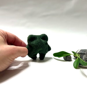 K&ouml;nnte beinhalten: Ein kleines, gr&uuml;nes Pl&uuml;schfroschspielzeug mit einer Hand, die sich nach ihm ausstreckt. Der Frosch steht auf einer wei&szlig;en Oberfl&auml;che mit einer kleinen gr&uuml;nen Pflanze und zwei grauen Steinen im Hintergrund.