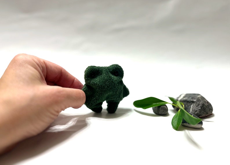 K&ouml;nnte beinhalten: Ein kleines, gr&uuml;nes Pl&uuml;schfroschspielzeug mit einer Hand, die sich nach ihm ausstreckt. Der Frosch steht auf einer wei&szlig;en Oberfl&auml;che mit einer kleinen gr&uuml;nen Pflanze und zwei grauen Steinen im Hintergrund.