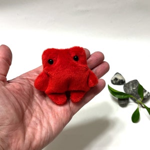 May include: A small, red, plush toy shaped like a creature with two black eyes. The toy is sitting in the palm of a hand.