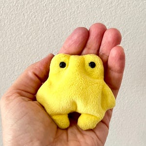 May include: A small, plush yellow creature with black button eyes and a soft, textured surface. The toy has a rounded body with small, stubby legs and arms. The toy is held in an open hand.