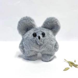 May include: A plush, gray creature with large, fluffy ears and a round body. The toy has black eyes and a small nose, giving it a cute appearance. The soft toy is standing on a white surface, with a few sprigs of dried grass nearby.