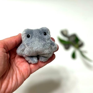 May include: A small gray plush frog with black eyes. The frog is being held in a person's hand.