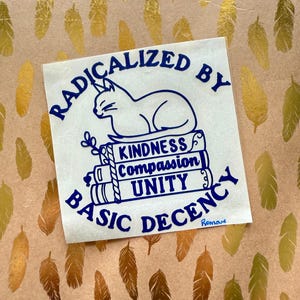 May include: A white sticker with blue text and a cat illustration. The text reads "RADICALIZED BY BASIC DECENCY" with the words "KINDNESS, COMPASSION, UNITY" on stacked books. The background has a gold feather pattern.