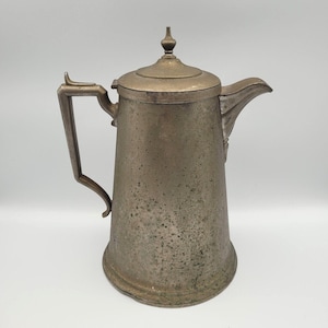 May include: An antique metal coffee pot with a lid and a spout. The pot has a handle and a textured, aged appearance. The color is a muted silver with darker areas, suggesting age and use. The design is simple and functional, typical of vintage kitchenware.