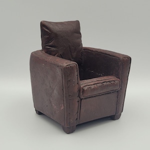 May include: Miniature brown leather armchair with a square back and a seat cushion. The small chair has a matching pillow and is a vintage style. The chair has a distressed look.