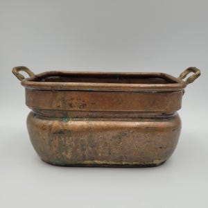 May include: A rectangular copper basin with rounded corners and two small brass handles on the sides. The metal surface shows a weathered, aged patina with visible oxidation and dark spots throughout the exterior of the container.