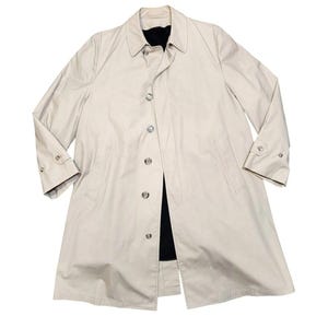 May include: A light beige trench coat with a button-up front, collar, and long sleeves. The coat has a classic design with a tailored fit, featuring buttoned cuffs and pockets. The interior lining is black. This coat is suitable for various occasions.