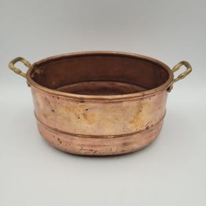 May include: A round copper basin with a polished finish and visible surface variations. The vessel features two small, curved brass handles attached to opposite sides of the rim. The exterior has a horizontal embossed line circling the middle.