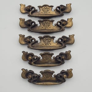 May include: Five antique brass drawer pulls with a decorative floral design. Each pull features a central emblem and curved handles. The pulls are arranged vertically against a neutral background, showcasing their aged, vintage aesthetic.