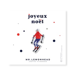 Op de afbeelding: Geborduurde strijkpatch met een skiër in een rood jack en blauwe broek, met de tekst "joyeux noël" in blauwe letters boven de skiër. De patch is op een witte achtergrond met kleine rode en grijze stippen.