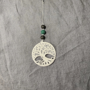 Puede incluir: Un charm de árbol de la vida de encaje blanco con cuentas negras y verdes, sujeto a un llavero plateado y una pulsera en espiral transparente. El charm es redondo con un diseño detallado de árbol.