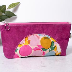 May include: A rectangular zippered pouch featuring a solid magenta suede-like exterior with a semi-circular front panel displaying a colorful fruit pattern of oranges, pink circles, and green leaves. A silver metal ring is attached to the zipper pull.