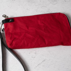 May include: A bright red clutch with a black leather wrist strap and silver-tone zipper. The rectangular bag is made of a textured fabric and features a black leather accent near the zipper.