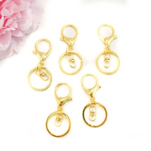 May include: Five gold-toned keychains with a lobster clasp and a round ring. The keychains are on a white background.