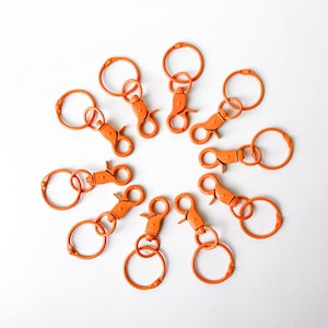 May include: A collection of orange keychains with lobster claw clasps and circular key rings. The keychains are arranged in a circular pattern against a white background. The key rings are approximately 1 inch in diameter.