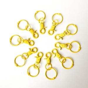 May include: Ten yellow metal keychains with a swivel clasp and a round ring. The keychains are arranged in a circle on a white background.