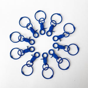 May include: A collection of ten cobalt blue keychains with lobster claw clasps and circular key rings. The keychains are arranged in a circular pattern against a white background. The key rings are approximately 1 inch in diameter.