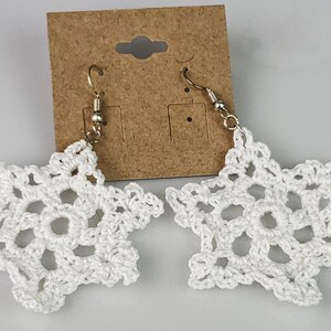 May include: A pair of white crocheted snowflake earrings with silver hooks. The earrings are displayed on a brown card.