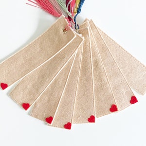 May include: Set of seven beige felt bookmarks with red heart accents. Each bookmark has a white border and a small heart at the bottom.