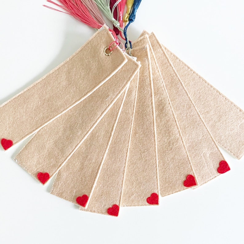 May include: Set of seven beige felt bookmarks with red heart accents. Each bookmark has a white border and a small heart at the bottom.