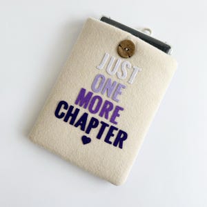 May include: A beige felt phone case with the text "JUST ONE MORE CHAPTER" in white, purple, and navy blue. A small navy blue heart is below the text. The case has a wooden button closure.