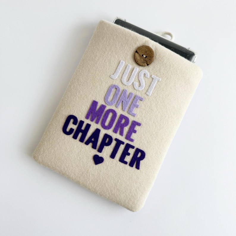 May include: A beige felt phone case with the text "JUST ONE MORE CHAPTER" in white, purple, and navy blue. A small navy blue heart is below the text. The case has a wooden button closure.