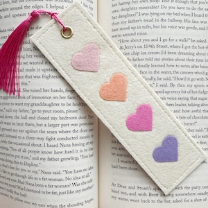 May include: A rectangular off-white felt bookmark featuring four small felt hearts in light pink, peach, bright pink, and lavender arranged vertically. The bookmark has a stitched border, a gold-toned metal eyelet, and a bright pink tassel.