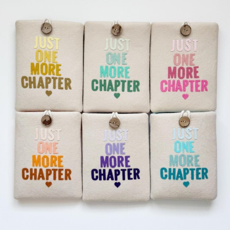 May include: Six beige book sleeves with the embroidered text "JUST ONE MORE CHAPTER" in various colors. Each sleeve has a wooden button closure and a small heart or heart-shaped design below the text.