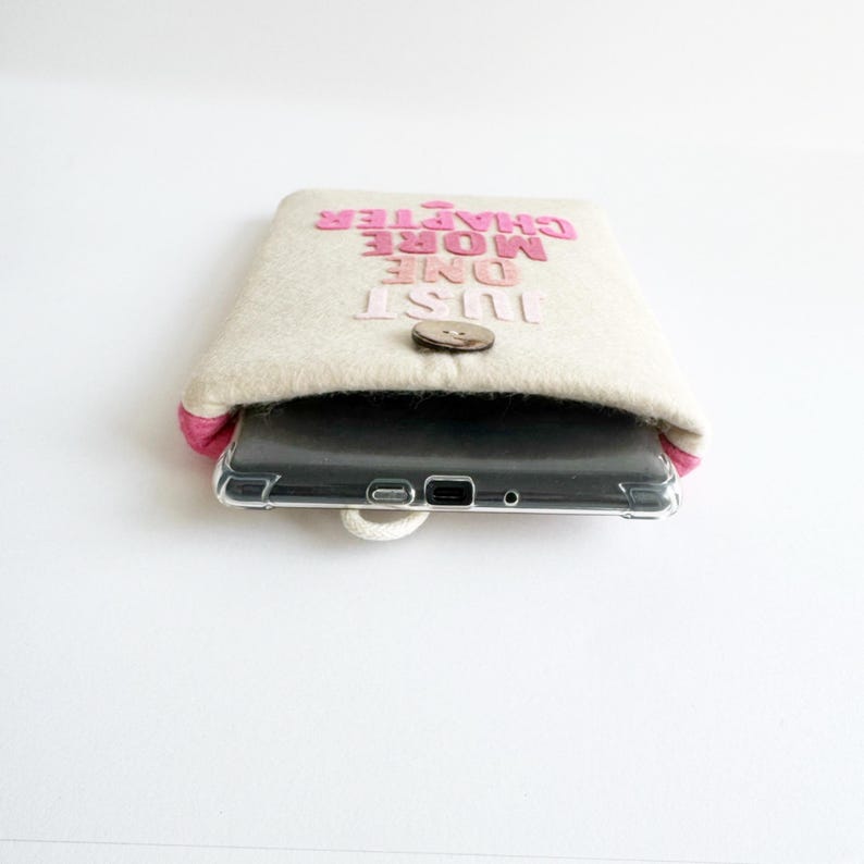 May include: Beige felt tablet sleeve with pink trim and the text "JUST ONE MORE CHAPTER" in pink. The sleeve has a wooden button closure and holds a clear phone case. The background is white.