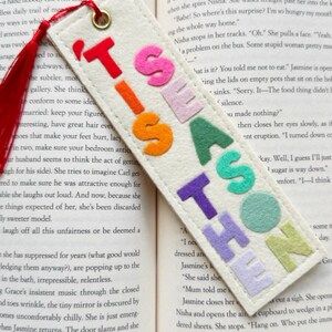 May include: Cream-colored felt bookmark with the text "TIS THE SEASON" in colorful, raised letters. The bookmark has a red tassel and a gold-colored grommet for attaching to a book.
