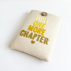 May include: A beige felt book sleeve with the text "JUST ONE MORE CHAPTER" in yellow and gold letters. A small gold heart is below the text. The sleeve has a wooden button closure at the top.