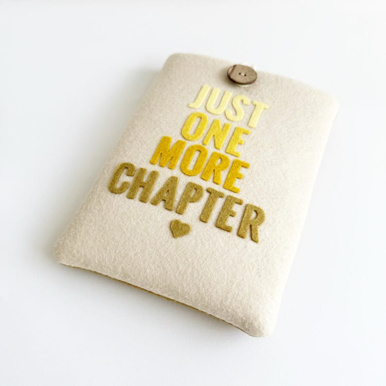 May include: A beige felt book sleeve with the text "JUST ONE MORE CHAPTER" in yellow and gold letters. A small gold heart is below the text. The sleeve has a wooden button closure at the top.