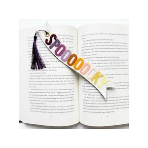May include: A white felt bookmark featuring the word SPOOOOKY in gradient-colored felt letters ranging from dark purple to yellow. The bookmark has a gold-toned metal eyelet at the top with a dark purple tassel attached, resting on an open book.