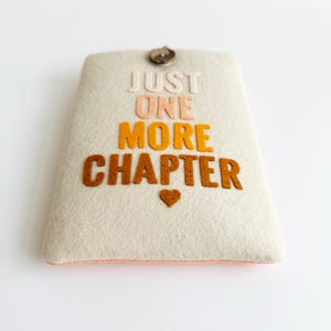 May include: A beige felt book sleeve with the text "JUST ONE MORE CHAPTER" in various colors. A small brown heart is below the text. The sleeve has a button closure at the top.