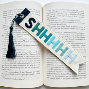 May include: A cream-colored felt bookmark featuring the word SHHHHH in a gradient of blue letters, ranging from dark navy to light sky blue. The bookmark includes a dark navy blue metal eyelet and a matching dark navy blue fabric tassel.