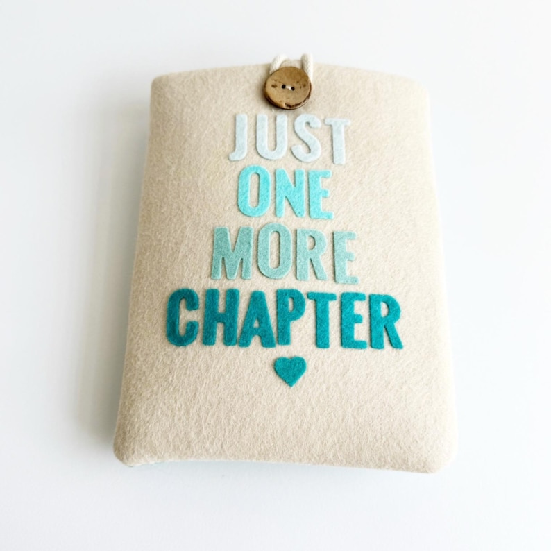 May include: A beige felt book sleeve with the text "JUST ONE MORE CHAPTER" in shades of blue. A small blue heart is below the text. The sleeve has a wooden button closure with a white cord loop.