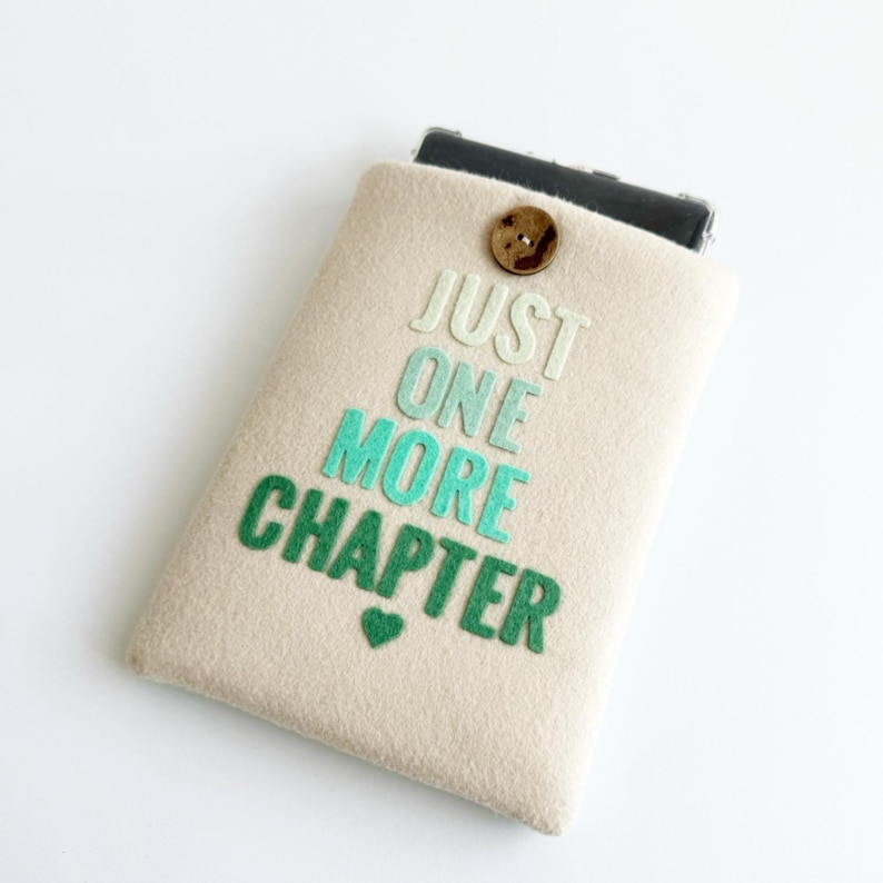May include: A beige felt phone case with the text "JUST ONE MORE CHAPTER" in shades of green and a small green heart. The case has a wooden button closure and holds a black phone.