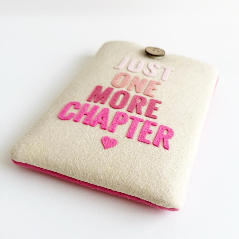 May include: A cream-colored felt tablet case with the phrase "JUST ONE MORE CHAPTER" in pink letters. A small pink heart is below the text. The case has a pink border and a wooden button closure.