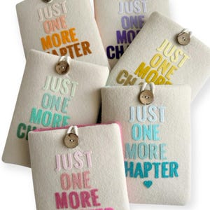 May include: A collection of off-white felt book sleeves, each embroidered with the phrase "JUST ONE MORE CHAPTER" in various colors. Each sleeve has a wooden button closure. The text colors include orange, purple, yellow, teal, and pink.