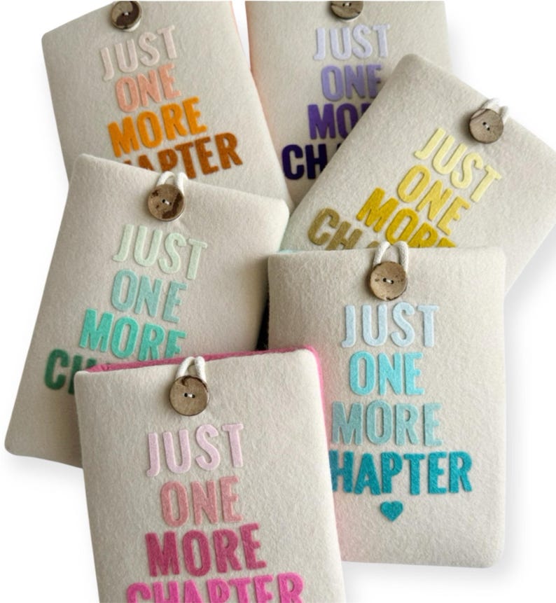 May include: A collection of off-white felt book sleeves, each embroidered with the phrase "JUST ONE MORE CHAPTER" in various colors. Each sleeve has a wooden button closure. The text colors include orange, purple, yellow, teal, and pink.