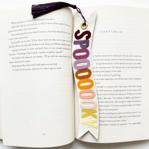 May include: A cream-colored felt bookmark with a pennant shape, featuring the word SPOOOOOKY in gradient felt letters ranging from dark purple to yellow. It includes a brass eyelet at the top with a dark purple tassel attached to the bookmark.