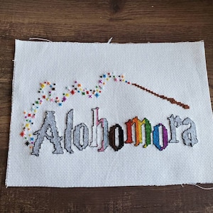 May include: White cross-stitch embroidery with the word "Alohomora" in rainbow colors, a wand, and a trail of colorful stars.