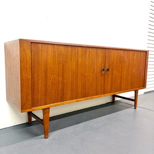 May include: A mid-century modern wooden cabinet with two doors and a warm brown finish. The cabinet has a rectangular shape, with a long, horizontal design. It features two round, brass-colored handles and is supported by angled wooden legs.