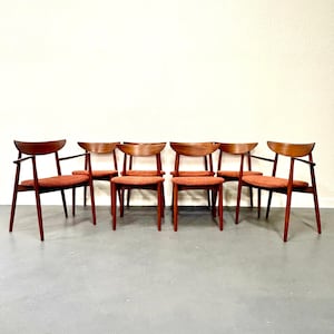 May include: A set of six mid-century modern chairs. The chairs have a dark wood frame with a curved back and armrests. The seats are upholstered in a reddish-orange fabric. The chairs are arranged in a row against a white wall.