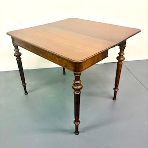 May include: A square wooden table with a rich brown finish and a visible wood grain pattern. The table features four slender, turned legs with decorative bulbous detailing and a smooth, flat top surface with a central seam line across the middle.