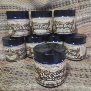 May include: A collection of eight jars of Back Forty Tallow Cream. The jars have a dark blue lid and a label with a landscape illustration and the text "Back Forty Tallow Cream". The label also lists ingredients.