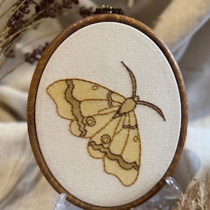 May include: A round, wooden embroidery hoop featuring a detailed moth design. The moth is embroidered in shades of yellow and brown on a cream-colored fabric. The wooden hoop has a natural finish, and the artwork is displayed on a clear stand.