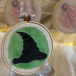 Op de afbeelding: Een kleine, ronde borduurring met een zwarte heksenhoed op een groene achtergrond. De hoed is gestikt met getextureerd garen. De ring wordt tentoongesteld op een heldere standaard, met andere soortgelijke ringen op de achtergrond.