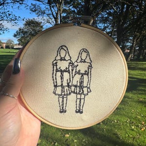 May include: An embroidery hoop featuring a black stitched design of two figures in dresses. The figures are side-by-side, with their arms touching. The embroidery is on a beige fabric, and the hoop is made of wood. The background is a grassy area with trees.