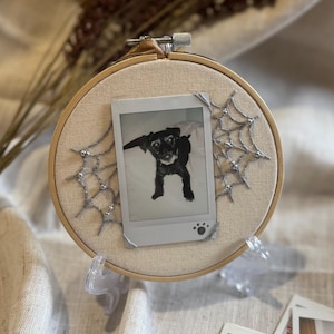 Peut inclure: Un cercle à broder rond avec un tissu beige en arrière-plan, présentant une photo Polaroid en noir et blanc d'un chien. Des décorations argentées en forme de toile d'araignée et un support transparent soutiennent le cercle. La photo a une empreinte de patte.